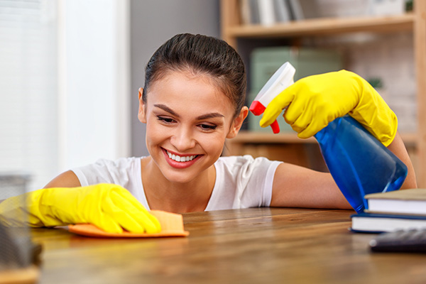 Smiling woman cleaning the desk with a rag while holding a spray bottle